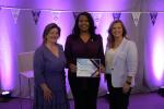 Three people standing together with a purple background, one holding a certificate