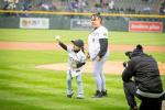 A child throwing a pitch on a baseball field, with an adult behind them and a photographer shown