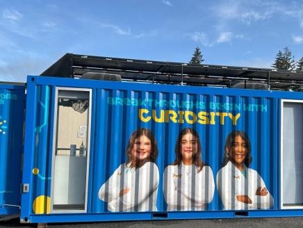 lays three The Curiosity Cube, a blue mobile science lab that displays three students in white lab coats and text that reads "Breakthroughs Begin With Curiosity"
