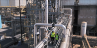 Two people wearing hard hats and high-vis vests walking on a long elevated pathway. Industrial piping and building behind them.