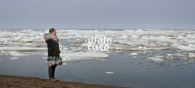 A person in warm clothing standing on a shore looking out over open water with chunks of ice and snow floating in it. "Cisco" overlaid.