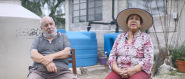Gloria and Sergio sitting in chairs outside, in front of large water collection containers.