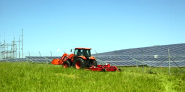 Large red tractor mowing a field next to a solar farm.