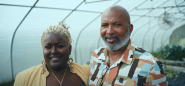 Two people standing in a greenhouse, an arm around each other.