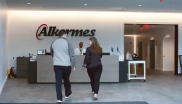 Two people approach a third at a reception desk. A sign "Alkermes" on the wall behind.