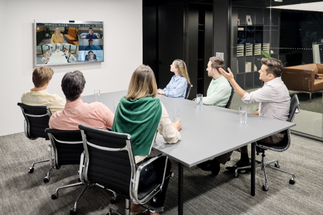 employees seated around a conference table viewing other team members on a monitor mounted on a wall 