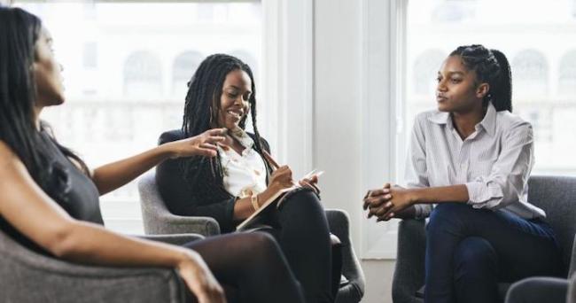 Three African American women in business attire are seated in a semi-circle having a discussion. The woman in the middle is taking notes on a pad. 