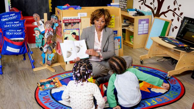 Black woman in business attire reading a book to young Black children