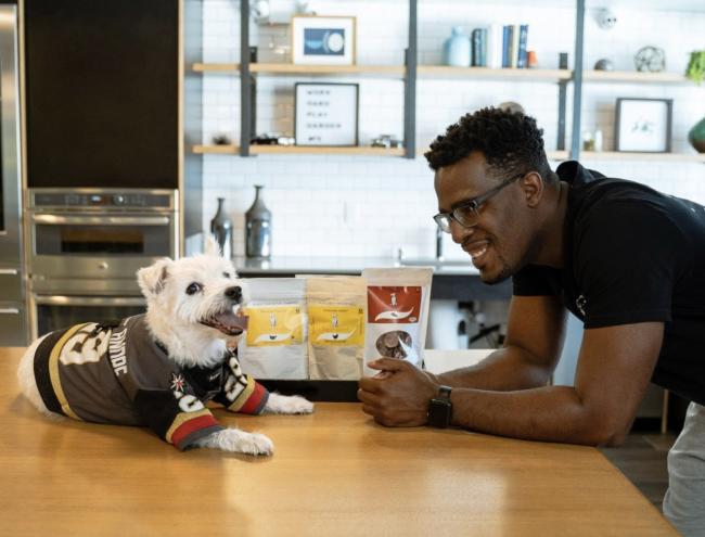 a small white dog in a sports jersey, sitting on a table with Eric Adams, in a black t-shirt, looking on