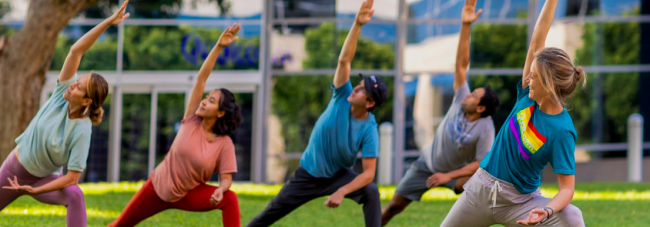 a group of 5 people do the same stretching exercise on a grass lawn outside of an office building