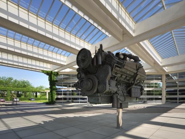 a large model motor on a stand in a cement walkway under glass and beam roof at Cumming headquarters
