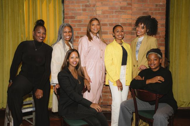 Queen Latifah and a group of others pose in front of a brick wall and yellow curtains