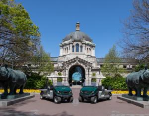 Two electric utility vehicles are parked in front of Bronx Zoo's Central Building on a sunny day. Flanking the vehicles are two animal statues.