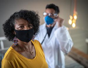 A medical professional using a stethoscope on a patient who is looking at the camera. Both are wearing protective masks.