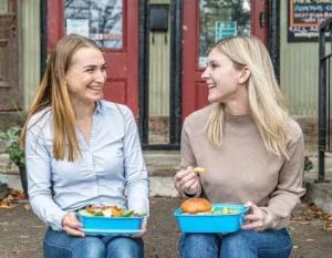 co-founders Jacquie Hutchings and Kayli Dale sitting on a front porch, eating out of blue containers
