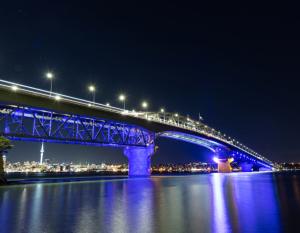 night panorama of Auckland Harbour Bridge over an expanse of water and is lit up in blue lights