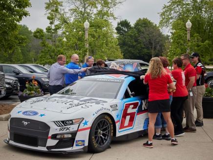 a group of people stand outside and take a cover off a race car. "6" on the side