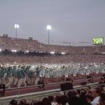 athletes dressed in matching uniforms walking along a track of a stadium. Stands filled with spectators.