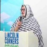 Faiza Yousuf standing at a podium, holding a microphone. Lincoln Corners Pakistan on the front