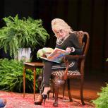 Dolly Parton sitting in a chair on stage reading a book, children in front of her