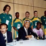 athletes in matching uniforms stand behind Nelson Mandela and other leaders seated at a long table