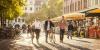 three people walk down a brick lined street, pedestrians and bike riders in the background on a sunny day